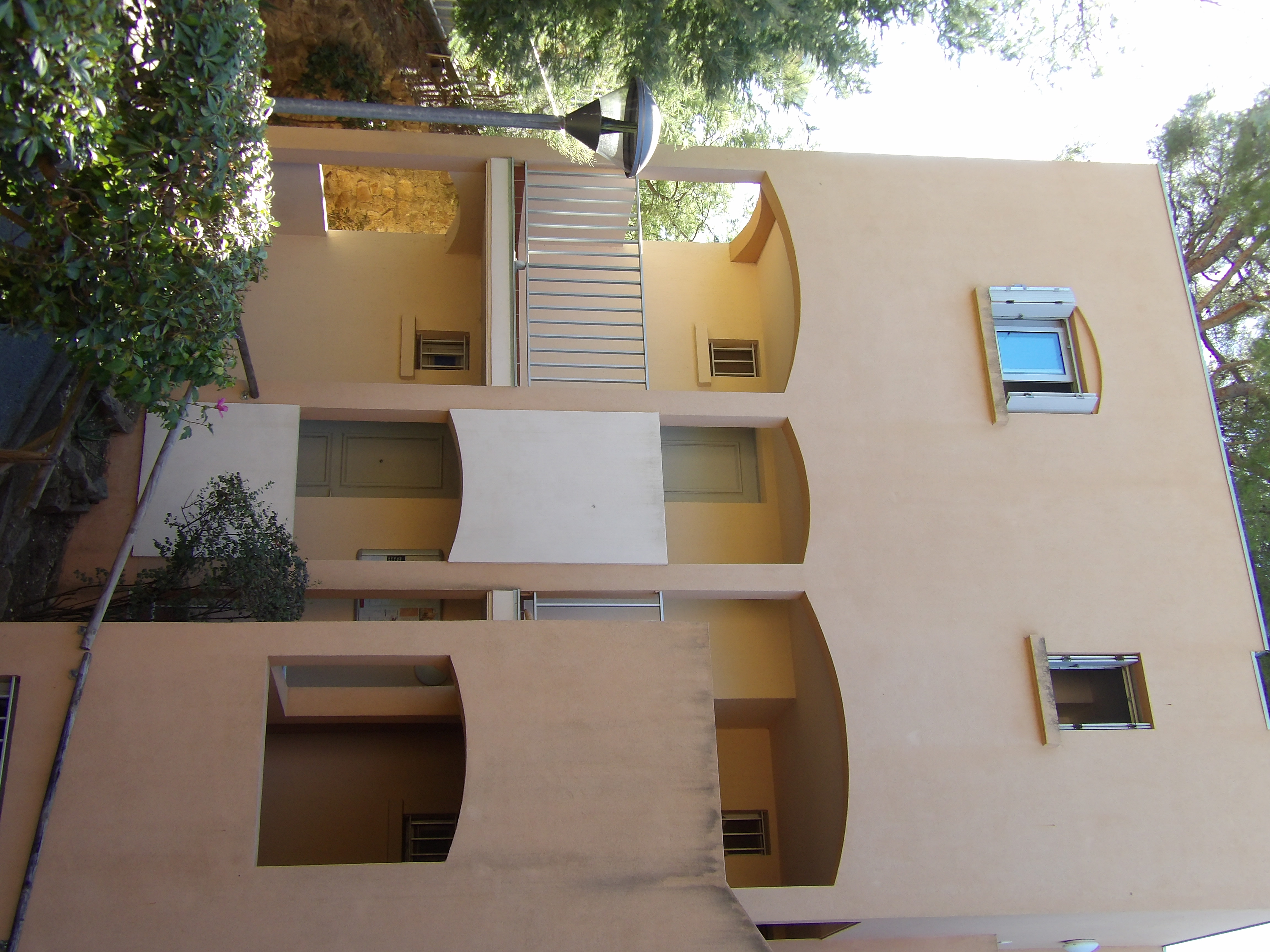 Façade extérieure de la résidence aux tons ocres, balcons et arches typiques de l'architecture provençale, entourée de pins
